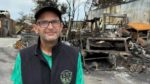 John Harvey stands in the destroyed yard of his MOT Centre. There are charred vehicles around him. He's wearing a dark cap, glasses. and a black gilet.