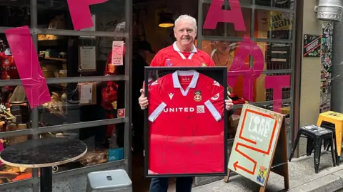Dieter Steinbusch Mike stood outside the pub holding a framed Wrexham shirt which has been signed.