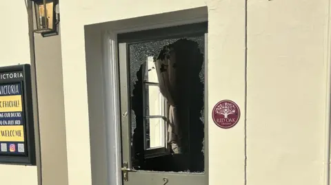 Finn Reeves A front door of a building with the window smashed. The door is light grey and the rest of the building is cream coloured