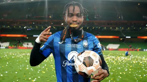 Ademola Lookman in blue and black Atalanta shirt stands on a pitch covered in glittering ticker tape after the 2024 Europa League final, holding the match ball, a trophy and with his medal between his teeth