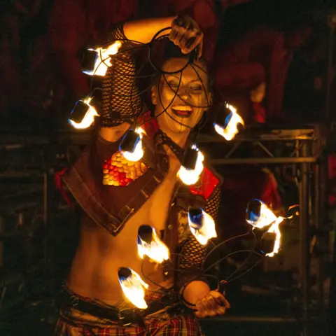 Zhi Kang Chua A man is smiling holding large metal rings that are on fire. he is wearing a black cropped denim top with fish net sleeves.