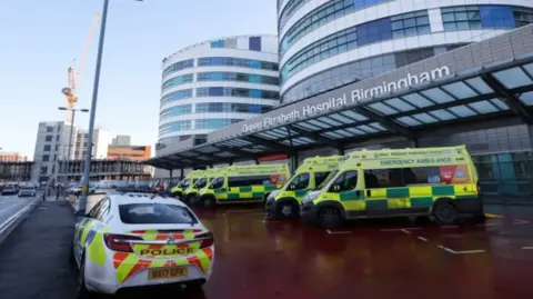 The front of a hospital building with a row of ambulances at the front and one police car. The sign above the door says Queen Elizabeth Hospital Birmingham
