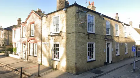 The Fountain is a pub situated on the corner where two roads meet. It is a brick building and has white framed windows and a gold sign on the left that says The Fountain.