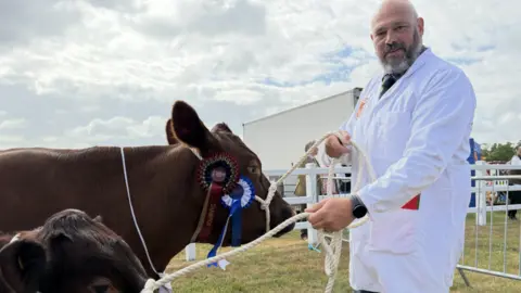 A man wearing a long white coat smiles at the camera. He holds two brown cattle to the left by ropes tied around their heads.
