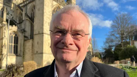 Jeremy Hilton smiles at the camera and stands outside a church on a bright sunny day. He has grey hair, glasses and wears a suit jacket