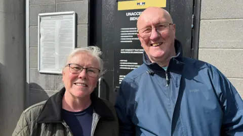 A man and a woman smile into the camera. They are standing outside a football stadium.