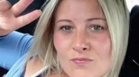 A woman with short blonde hair. She is wearing a blue top and pouting at the camera as she holds one hand up in a peace sign.