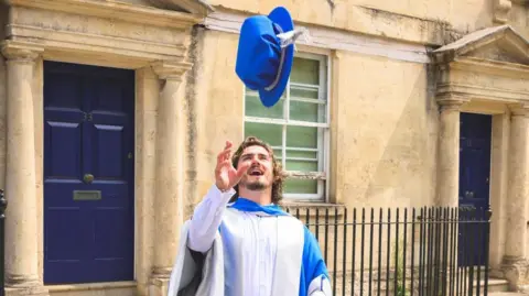 A man in a blue and white graduation gown throwing his graduation hat into the air as he smiles openly and watches his hat. His right arm is raised slightly as holds his hand open ready to catch the cap.
He is standing in front of a pavement and some Bath houses with dark doors. 