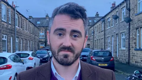 BBC A man with a dark beard and silver/grey hair wearing a brown suit jacket and purple V-necked sweater standing in a terraced street.