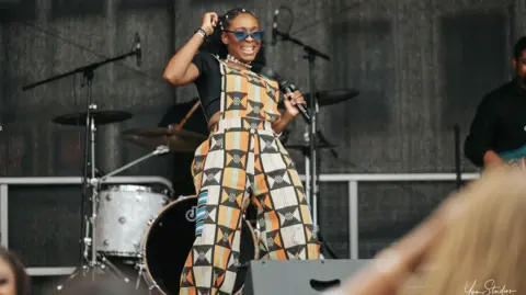Yom Studios Photography Woman performing on an outdoor stage, holding a microphone and wearing blue sunglasses and a patterned orange, black, and cream jumpsuit. A drum kit and microphone stands are in the background, with audience members blurred in the foreground.