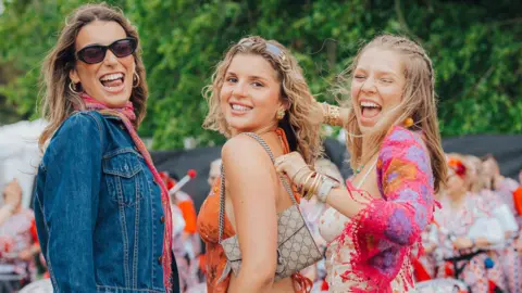 Jessie Myers Three young women turn to smile at the camera. They are wearing bright clothes, including halternecks and strappy tops.