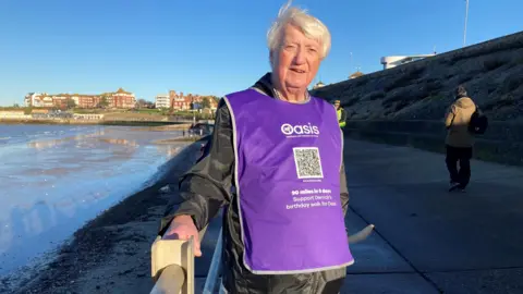 Derrick Downs looking to camera wearing a mauve bib standing beside the sea on the sea wall with buildings behind him in the distance in the sunshine