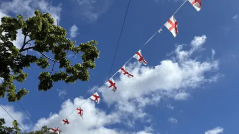 England flag bunting flies in a blue sky with a couple of clouds. A green leafy tree is visible in the top left of the picture.