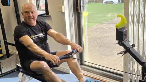 A man with short grey hair smiles as he uses a rowing machine. The rowing machine is positioned next to a patio door which looks out onto a paved concrete area and grass. He is wearing a black T-shirt with a white and green "Concept 2" logo on the chest, black shorts and bright blue trainers.