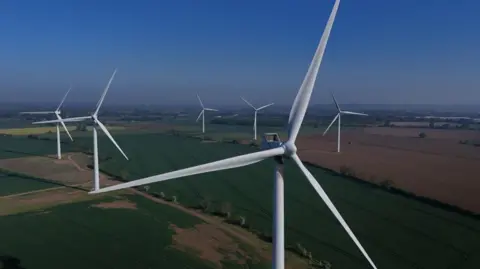 An aerial view of a wind farm in countryside. In the foreground is a very large white wind turbine. The tower supports three long blades. Several more wind turbines are arranged in across the fields. Low hedgerows separate some of the fields.