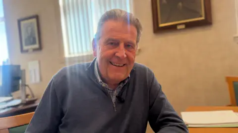 Man in his 60s seated at a desk inside a parish hall office, with papers on the table and framed pictures on the wall behind.