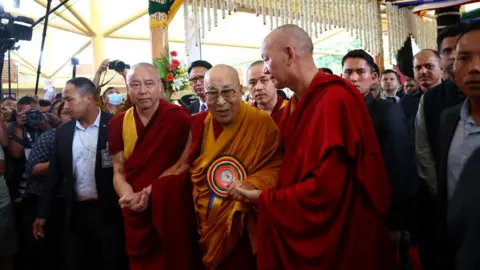 The bespectacled Dalai Lama cloaked in a red robe with a flowing orange wrap, arrives for his birthday celebrations flanked by two Buddhist's in red robes holding his hand. They are surrounded by suited men and a number of photographers.