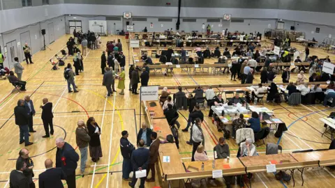 A sports hall with lots of tables in row and people milling around. Signs on the tables indicate geographical areas and identify this as an election count.
