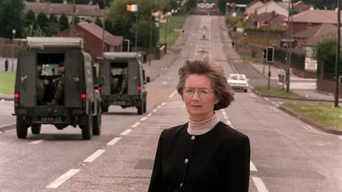 Pacemaker Bríd Rodgers standing on the Garvaghy Road in Portadown in 1998.  She is wearing a black jacket with gold buttons over a pale turtleneck top.  There are two armoured Army vehicles behind her.  The rear doors of both vehicles are open and soldiers holding guns are inside.  Irish tricolour flags are flying from lamp posts on the road behind her.