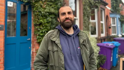 A man with brown hair and a beard is smiling as he stands outside the blue painted door of a red brick house which has plants on the walls. He is wearing an olive green coat and a blue hooded top. Purple and blue wheelie bins are behind him.