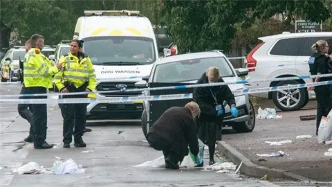 Ryan Underwood Police officers work at the scene of an incident. A section of the street is cordoned off with blue and white police tape. Some police are talking while others are looking at evidence on the ground. Cars can be seen parked beyond the cordon. 