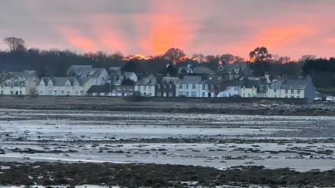 Graham Young Orange rays over houses in Wigtownshire. The rays are orange and on the background of a lilac-grey sky. The houses in the foreground are all different colours including pale yellow, brown and white.