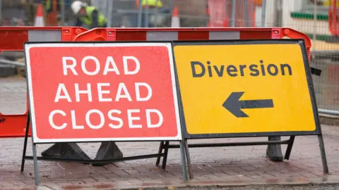 Getty Images A red sign with white writing reading "road closed ahead" in capital letters. On the right is a yellow sign with "diversion" written in black with a black arrow beneath it pointing to the left.