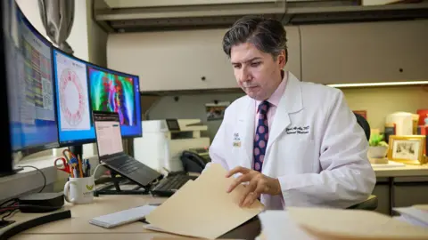 Ozempic and Wegovy might spice up well being, from habit to dementia Matt Miller/WashU Medicine Man in a pink shirt and tie with a white lab coat flips through medical reports in his office.