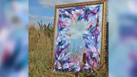 Sophie Tea Art The mirror photographed propped up in a grassy field with blue sky behind it. It's in a gilded metal frame and is painted around the edges with an abstract flower pattern in shades of blue, purple, pink and white.