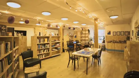 Ben Glasgow Inside the library there are books on shelves all around the room. There is a table in the middle and several chairs dotted around the room. There are a strip of lights along the ceiling because there are no windows as its in a basement.