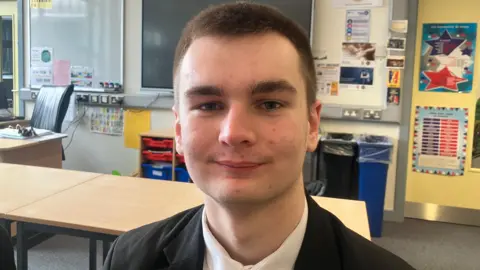 Adam, a teenage schoolboy, standing in a classroom and pictured from the shoulders up. He wearing a white shirt under a black blazer. He has short brown hair. Behind him there are desks, a blackboard and a whiteboard. 