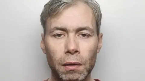 Thames Valley Police Liam Jones' mugshot. A middle aged man, with a greying beard and grey hair in front of a grey background