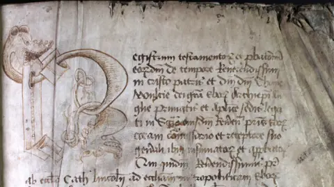 Borthwick Institute for Archives, University of York A paragraph of Latin text within a probate register, including a large letter R illustrated by two entwined dragons. The writing is clearly old, and the paper is worn and burned at the edges.