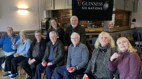 Seven people sit in chairs holding red balls. Two people stand behind them. They are pictured in a clubhouse which has a bar area behind them. 