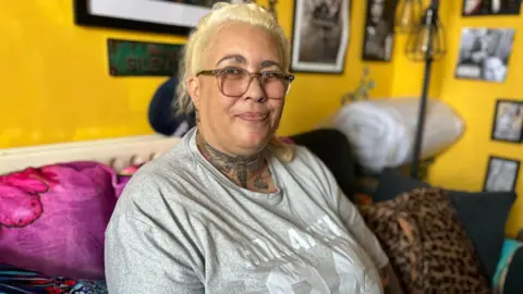 Dee is sat in her living room on a sofa, the wall behind her is yellow and there are framed pictures on the walls. She is smiling at the camera and wearing a grey printed t-shirt and glasses.