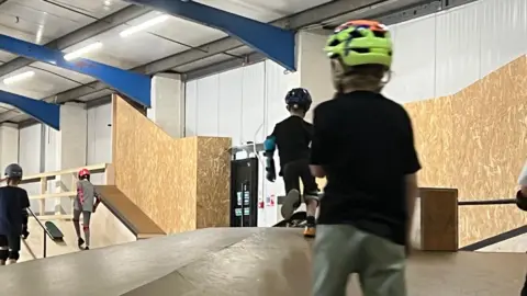 Mousumi Bakshi/BBC Several children are riding skateboards on a skatepark which is located in a white warehouse. The image is focusing on one child who is on the right and who is wearing a green and orange helmet, black T-shirt and green trousers.
