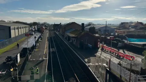 A railway line and station are pictured from a bridge. People in high-visibility vests stand on the platform. 