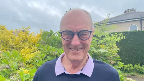 An image of Gordon Birtwistle in front of a green bush. He is smiling and wearing blue glasses, he is also wearing a navy jumper over a purple shirt