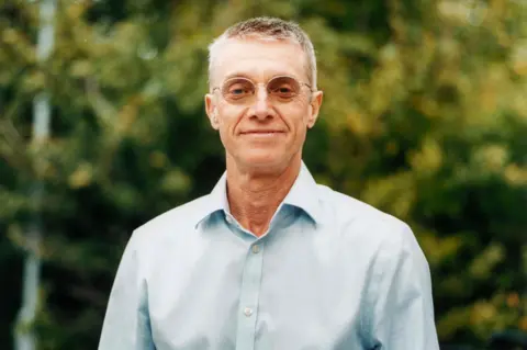 Francis Augusto Prof Sir Peter Horby wears a light blue shirt with the top button undone. He has short grey hair and small oval glassses.