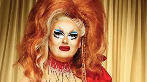 Drag artist Porkchop with a red dress on and wearing a large red wig with blonde streaks.