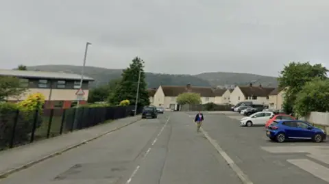 A street view, with parked cars lined up on the right, and a school on the left. A person is seen crossing the road