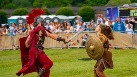 Chester Racecourse A man and woman dressed up in Roman armour stage a battle on a field at Chester Racecourse for Roman Day as crowds of families watch. 