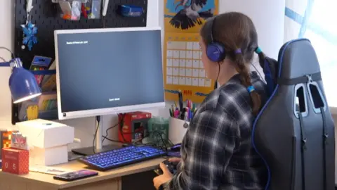 Ruby playing her game on a PC monitor. She has headphones on and is holding a controller while sitting on a chair in her bedroom.