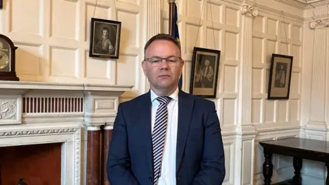 Philip White wears a suit and stripey tie, in the county building. He has short dark hair, is not smiling and stands in a white-panelled room.