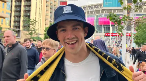 Dan Wolfendale is wearing a navy bucket hat and has a yellow scarf around his neck. He is smiling in front of a crowded Wembley Stadium.
