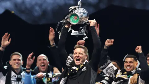 Eddie Howe lifting the Carabao cup above his head which is a silver cup with a green bottom. He has light brown, short hair and is wearing a black Newcastle United long-sleeved top and a scarf around his neck. He has his mouth open as if shouting and looks very happy. Newcastle players, Bruno Guimarães, Sven Botman and John Ruddy stand behind him smiling with their arms in the air. They are on a stage with a black background.