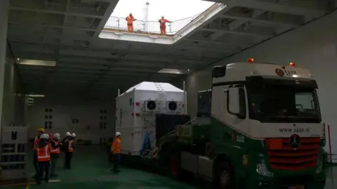 UCL A large, white container on the back of a lorry. It is inside a ship's cargo hold and is surrounded by workers in high-visibility clothing.