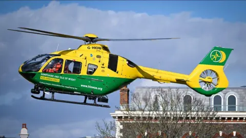 A yellow and green helicopter is pictured from the side, with its nose facing the left. It is in flight and a rooftop and the upper branches of a bare tree can be seen in the background. 