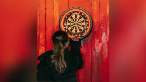 Factory Floor A woman with long dark blonde hair holds dart in front of a dartboard. The dartboard surround is orange and black and the dartboard is mounted on a wood panelling which is painted a rustic red. The woman is photographed from behind.