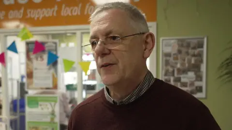 A man with grey hair who is wearing frameless glasses and a brown jumper over a checked short. He is standing in a community centre which has light green painted walls and a window with information posters on.